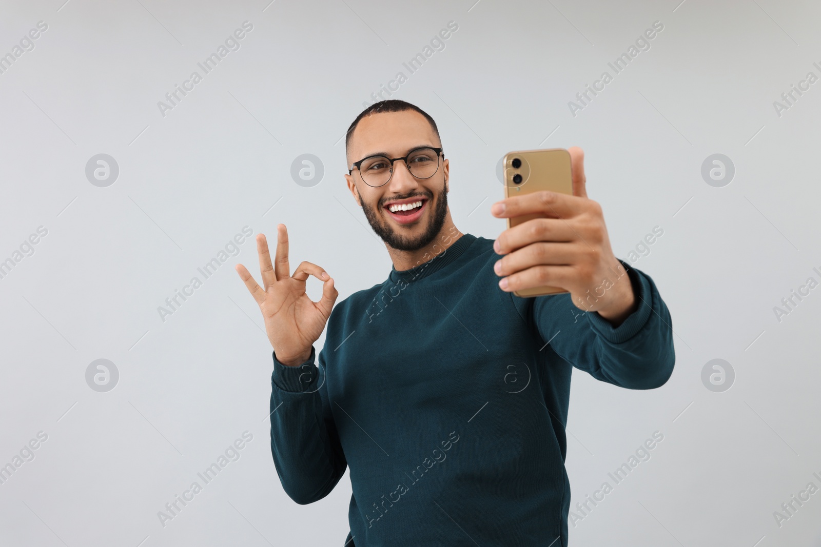 Photo of Smiling young man taking selfie with smartphone and showing OK gesture on grey background