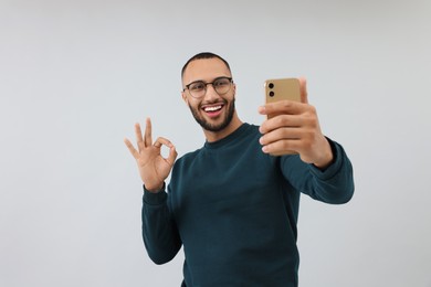 Smiling young man taking selfie with smartphone and showing OK gesture on grey background