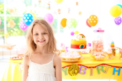 Cute little girl near table with treats at birthday party indoors