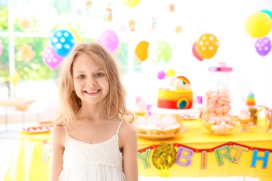 Photo of Cute little girl near table with treats at birthday party indoors