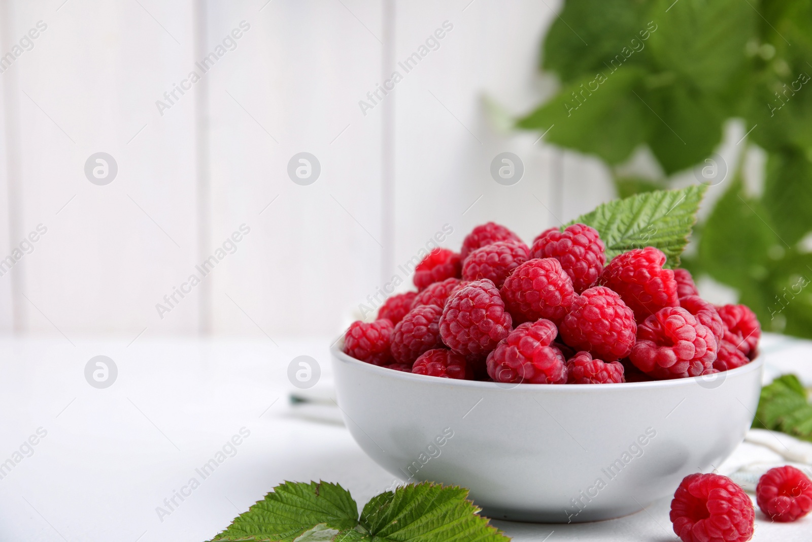 Photo of Bowl of fresh ripe raspberries with green leaves on white table against blurred background. Space for text