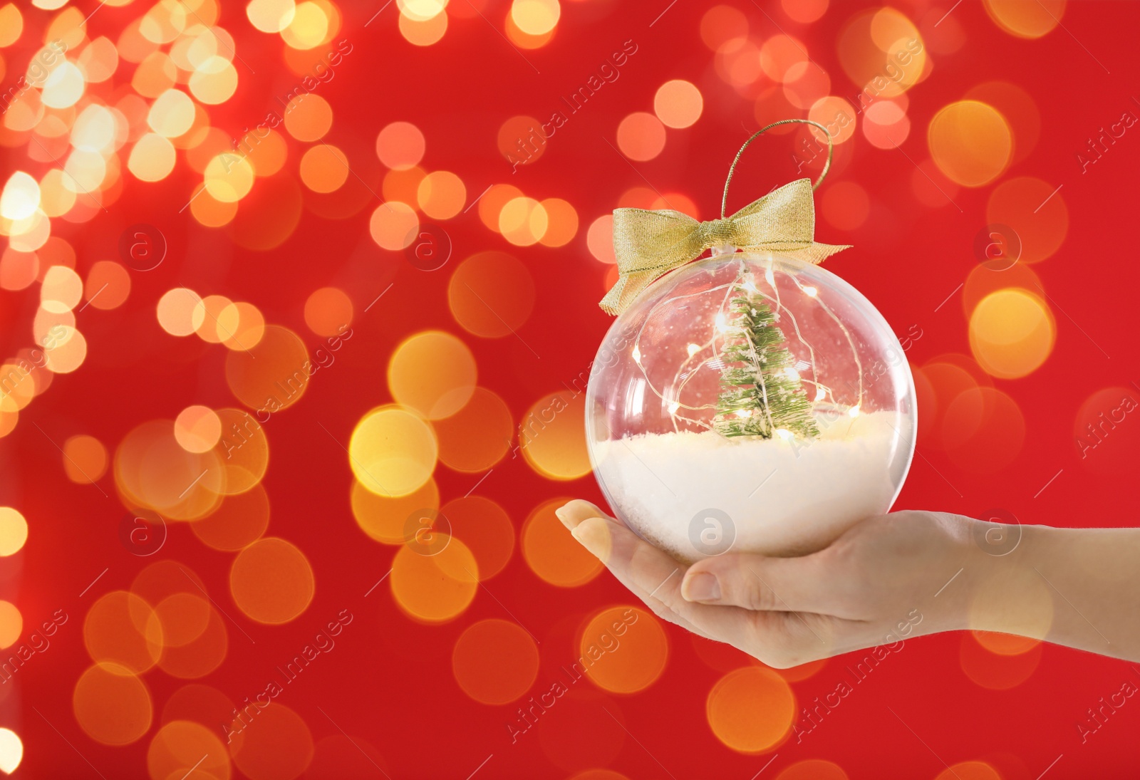 Image of Woman holding transparent Christmas ornament with small tree, fairy lights and snow against red background, bokeh effect. Space for text