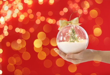 Woman holding transparent Christmas ornament with small tree, fairy lights and snow against red background, bokeh effect. Space for text