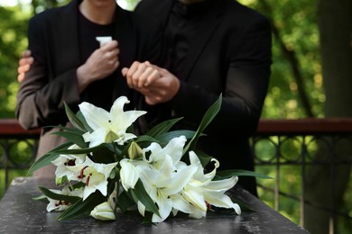 Couple near granite tombstone with white lilies at cemetery outdoors, selective focus. Funeral ceremony