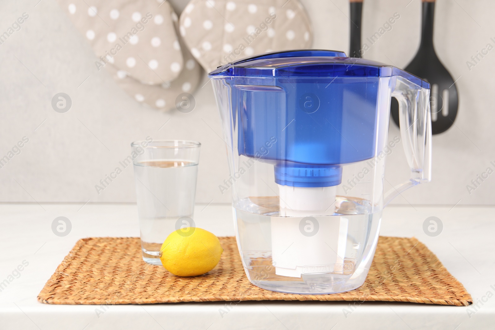 Photo of Water filter jug, glass and lemon on white countertop in kitchen