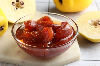 Photo of Tasty homemade quince jam in bowl and fruits on tiled table, closeup