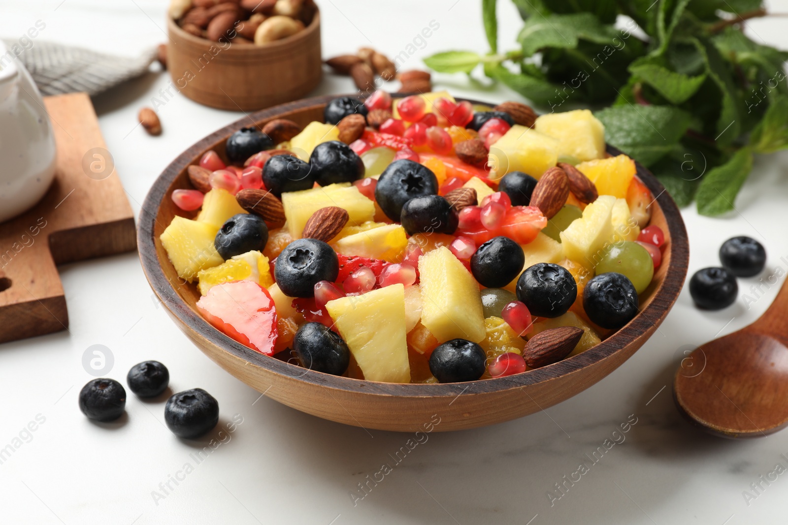 Photo of Delicious fruit salad in bowl, berries, fresh mint and nuts on white marble table
