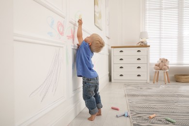 Photo of Mischievous little boy drawing with colorful chalk on white wall at home