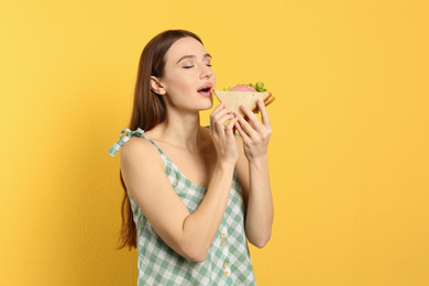 Young woman eating tasty sandwich on yellow background