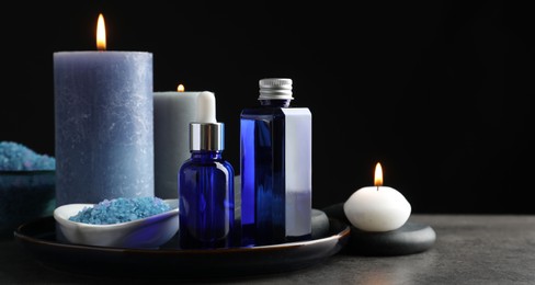 Photo of Spa composition. Cosmetic products, burning candles and sea salt on gray table against black background, space for text