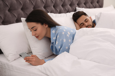 Photo of Young woman using smartphone while her boyfriend sleeping in bed at home