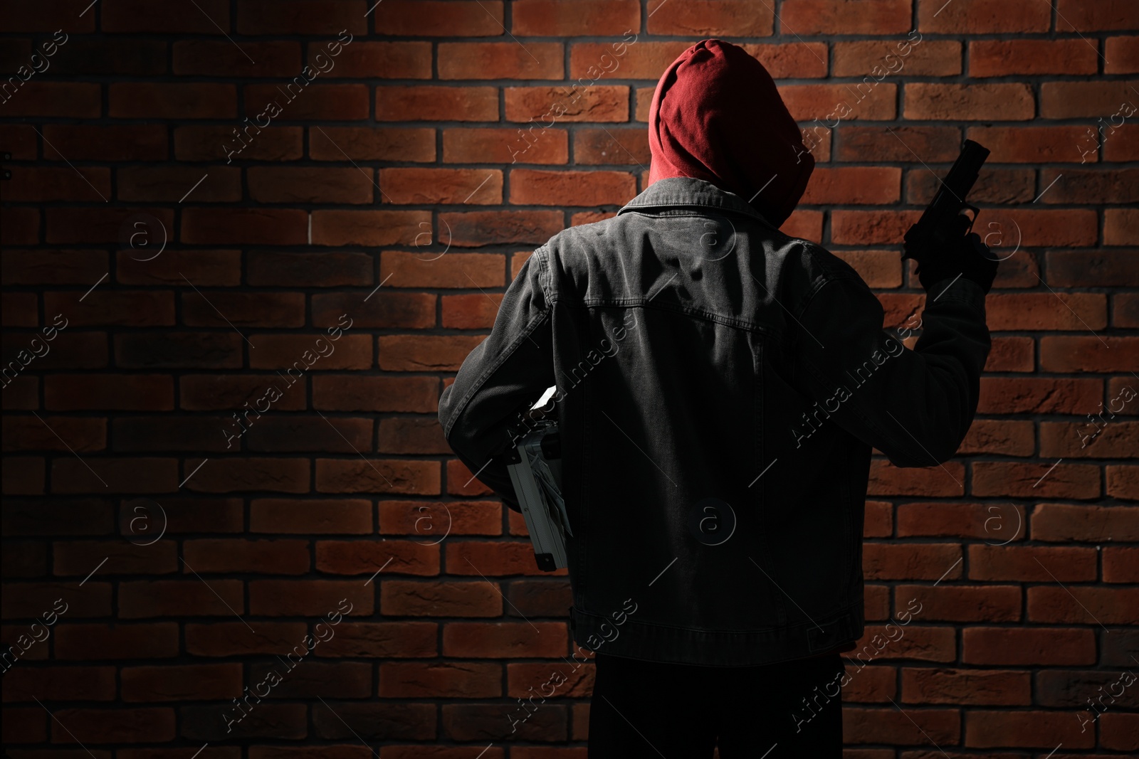 Photo of Thief in hoodie with gun against red brick wall, back view. Space for text