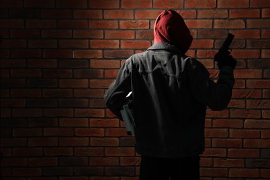 Photo of Thief in hoodie with gun against red brick wall, back view. Space for text