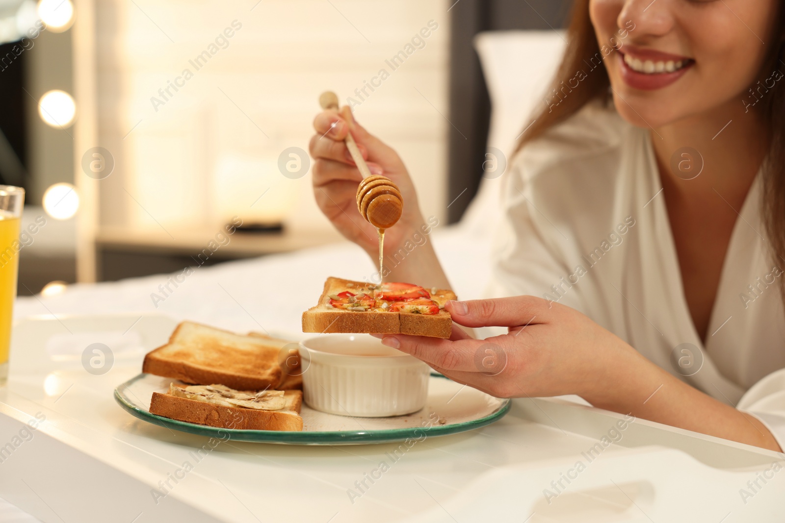 Photo of Happy young woman pouring honey onto toast near white tray on bed at home, closeup