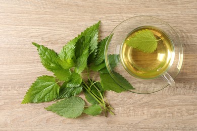 Glass cup of aromatic nettle tea and green leaves on wooden table, flat lay
