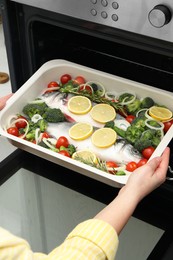 Photo of Woman putting baking dish with raw fish and vegetables into oven, closeup