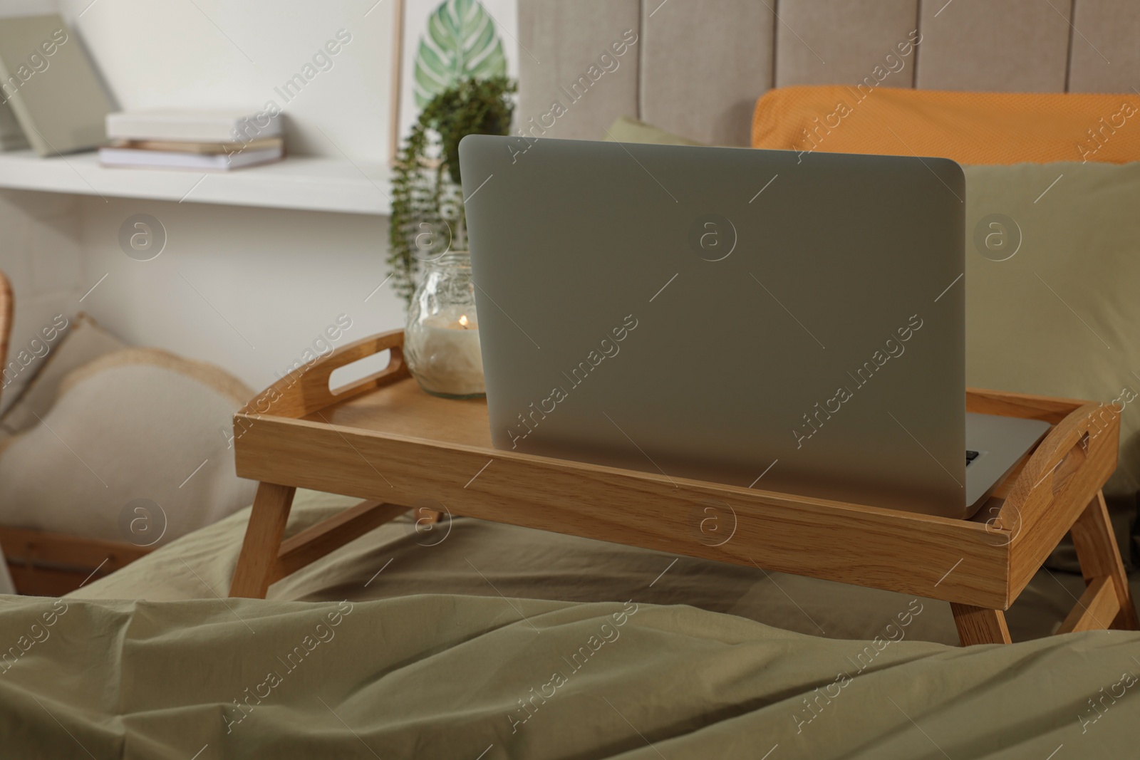 Photo of Wooden tray with modern laptop and burning candle on bed indoors