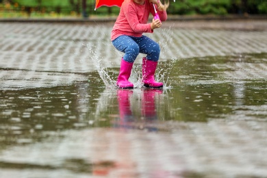 Photo of Little girl jumping in rubber boots under rain on street, closeup