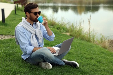 Man in sunglasses with laptop and coffee on green grass near lake