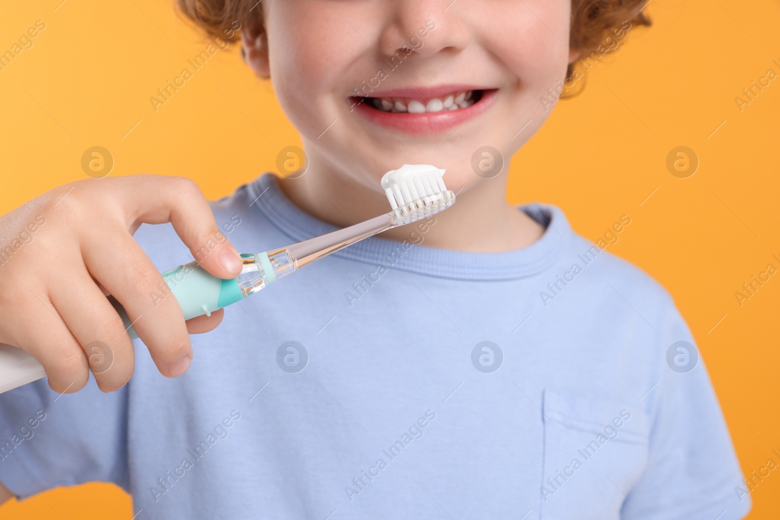 Photo of Cute little boy holding electric toothbrush with paste on yellow background, closeup