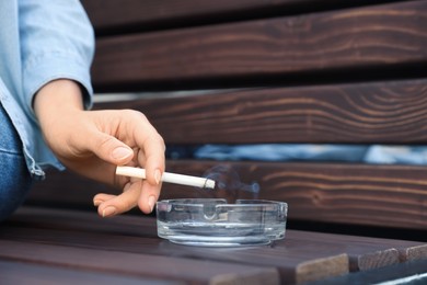 Photo of Woman holding cigarette over glass ashtray on bench outdoors, closeup