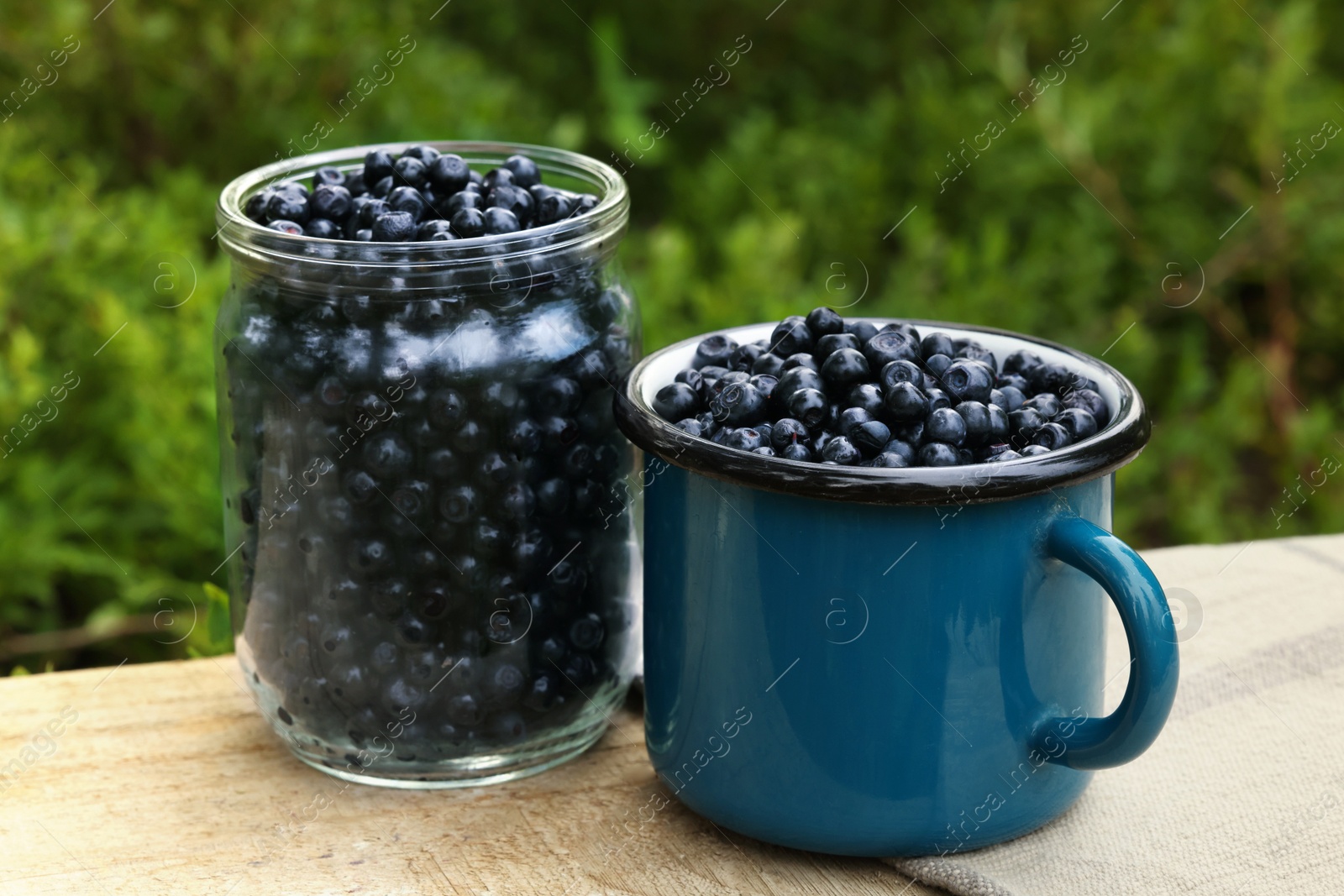 Photo of Jar and cup of delicious bilberries on wooden table outdoors, closeup