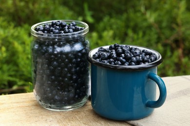 Photo of Jar and cup of delicious bilberries on wooden table outdoors, closeup