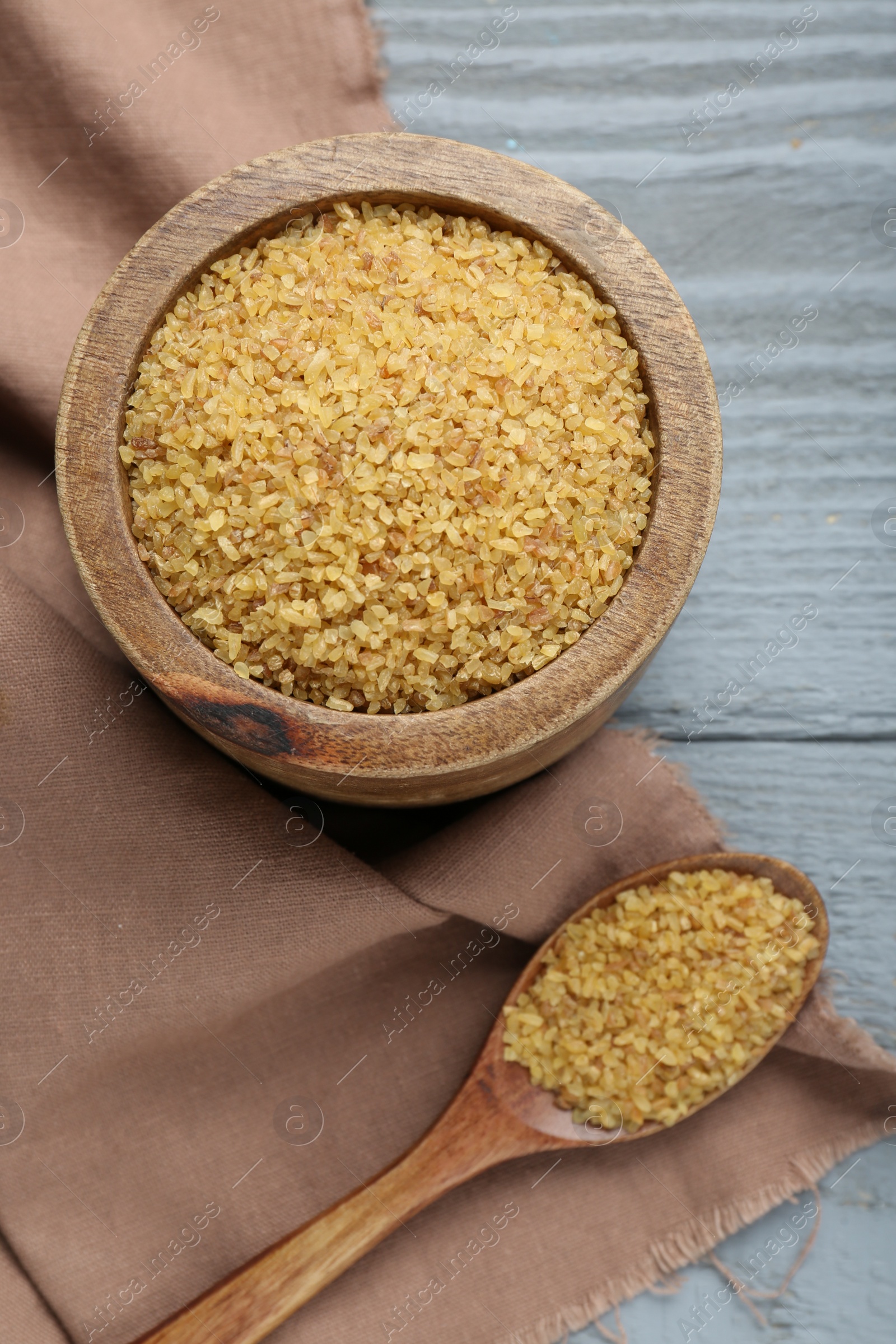 Photo of Bowl and spoon with uncooked bulgur on grey wooden table, flat lay