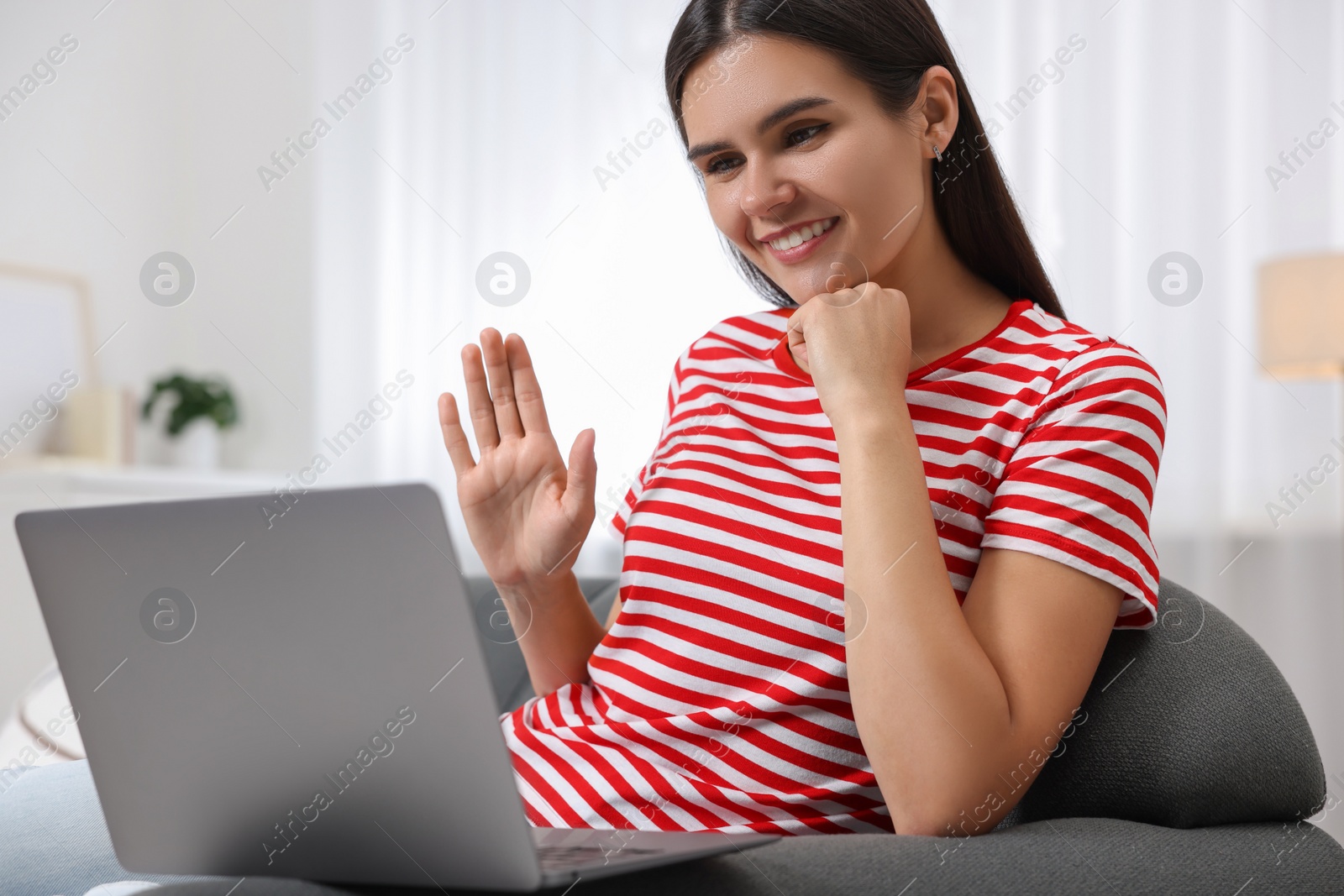 Photo of Happy young woman having video chat via laptop and waving hello on sofa in living room