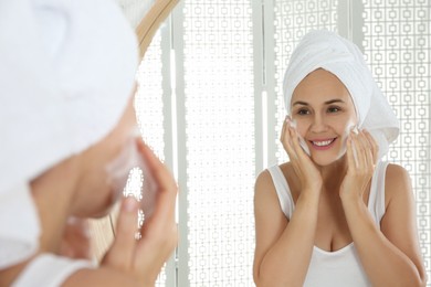 Photo of Happy mature woman applying cleansing foam onto face near mirror in bathroom