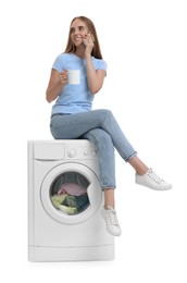 Photo of Beautiful young woman with cup of drink talking by smartphone on washing machine with laundry against white background