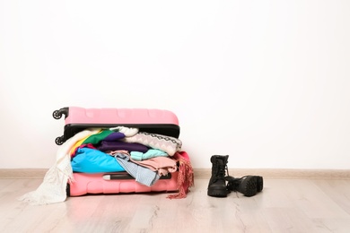 Photo of Suitcase with clothes and boots on floor against white wall, space for text. Winter vacation