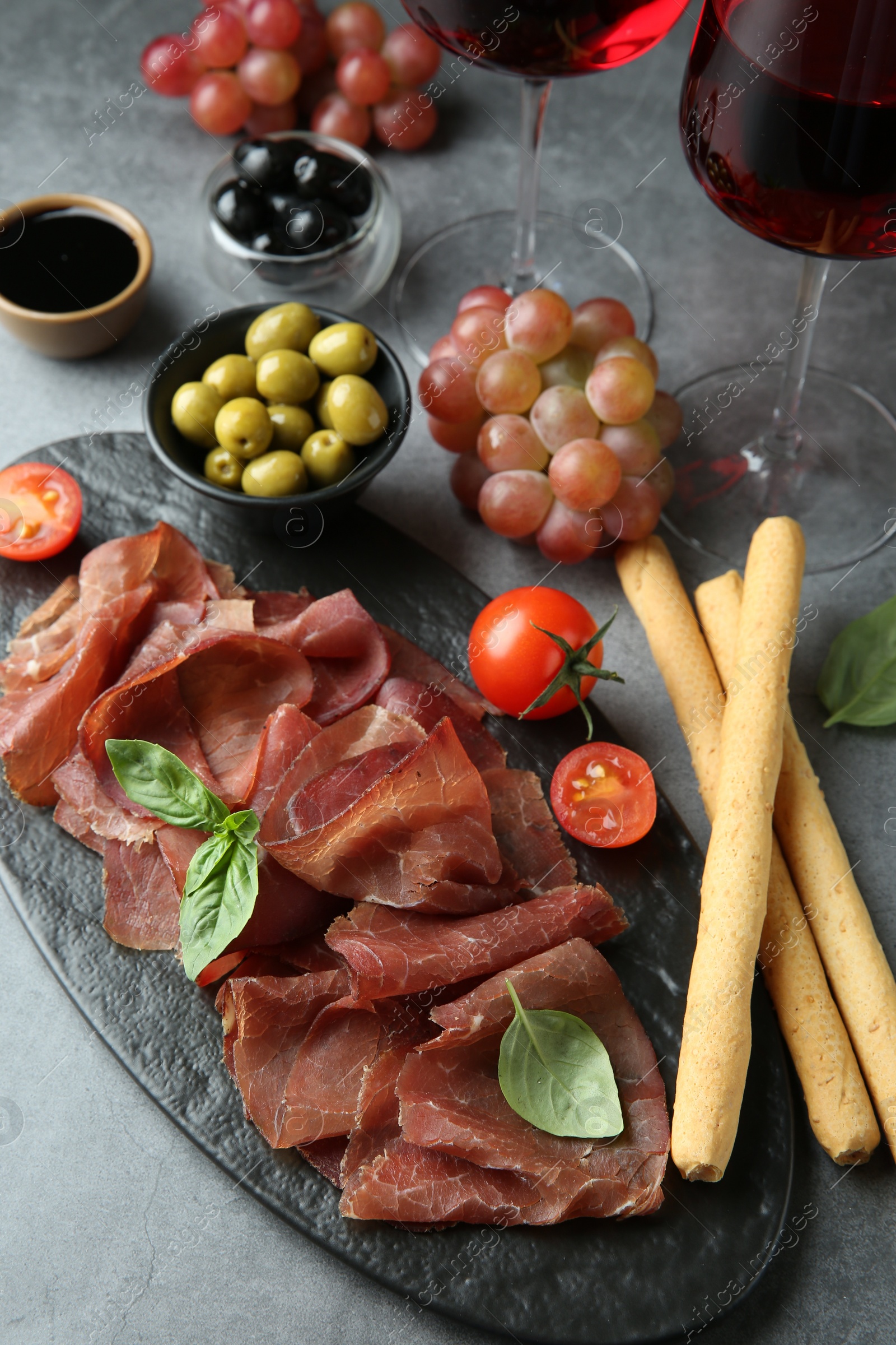 Photo of Delicious bresaola, grissini sticks, olives, tomato and grapes on grey textured table, above view