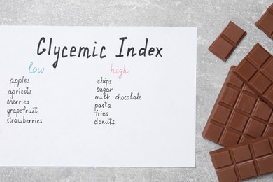 Photo of List with products of low and high glycemic index and chocolate on light grey table, top view