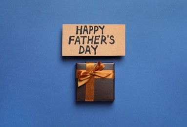Photo of Card with phrase Happy Father's Day and gift box on blue background, flat lay