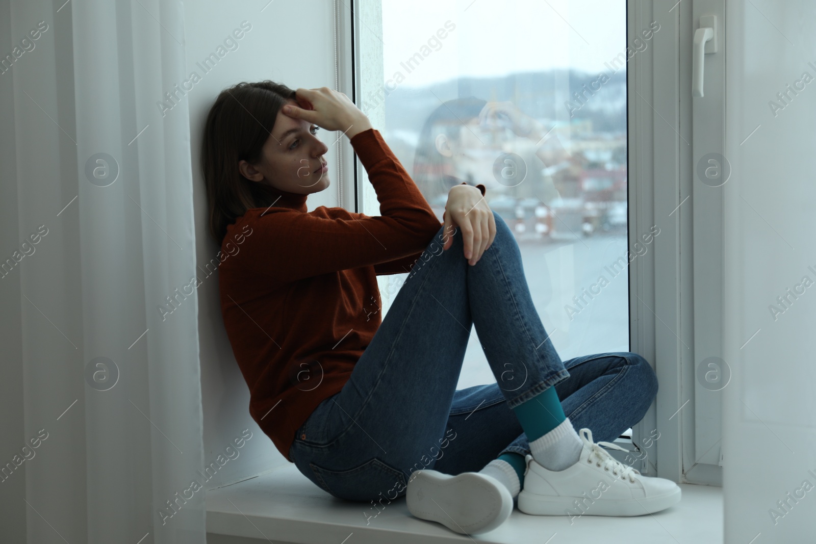 Photo of Sad young woman sitting on windowsill near window at home