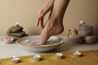 Photo of Woman soaking her foot in bowl with water and petals on white wooden floor, closeup. Pedicure procedure