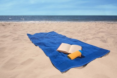 Photo of Soft blue beach towel, sunblock and book on sandy seashore