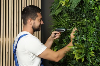 Photo of Man with screwdriver installing green artificial plant panel on grey wall in room
