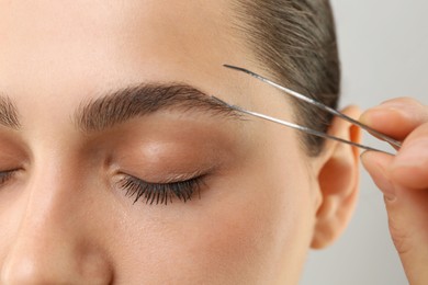 Photo of Eyebrow correction. Young woman with tweezers on light grey background, closeup