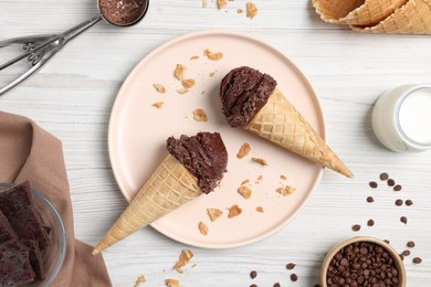 Chocolate ice cream scoops in wafer cones, milk and candies on light wooden table, flat lay