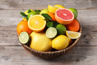 Photo of Different fresh citrus fruits and leaves in wicker basket on wooden table, closeup