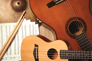 Photo of Ukulele, acoustic guitar, drumsticks, cymbal and note sheets on wooden background, flat lay. Musical instruments
