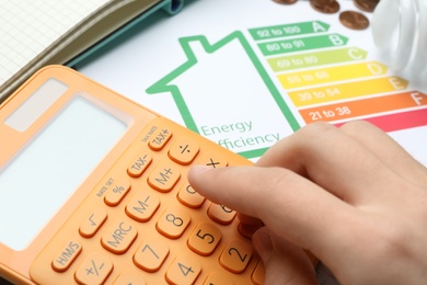 Photo of Woman with calculator and energy efficiency rating chart at table, closeup