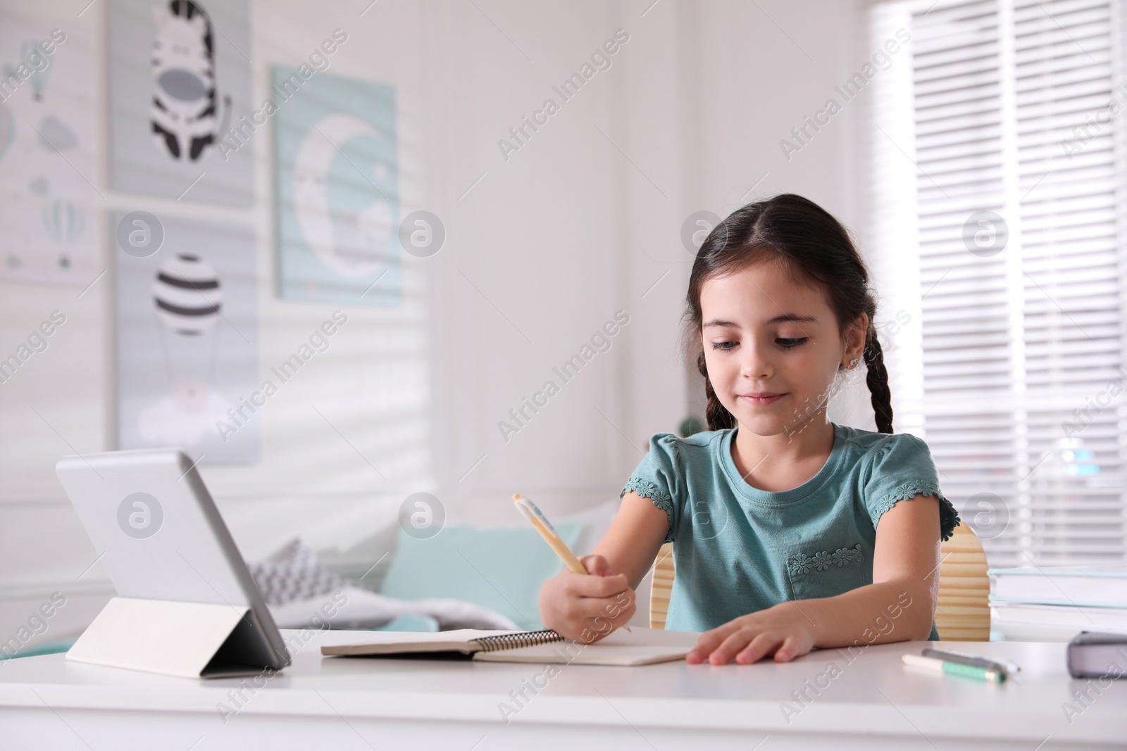 Photo of Little girl doing homework with tablet at table in room