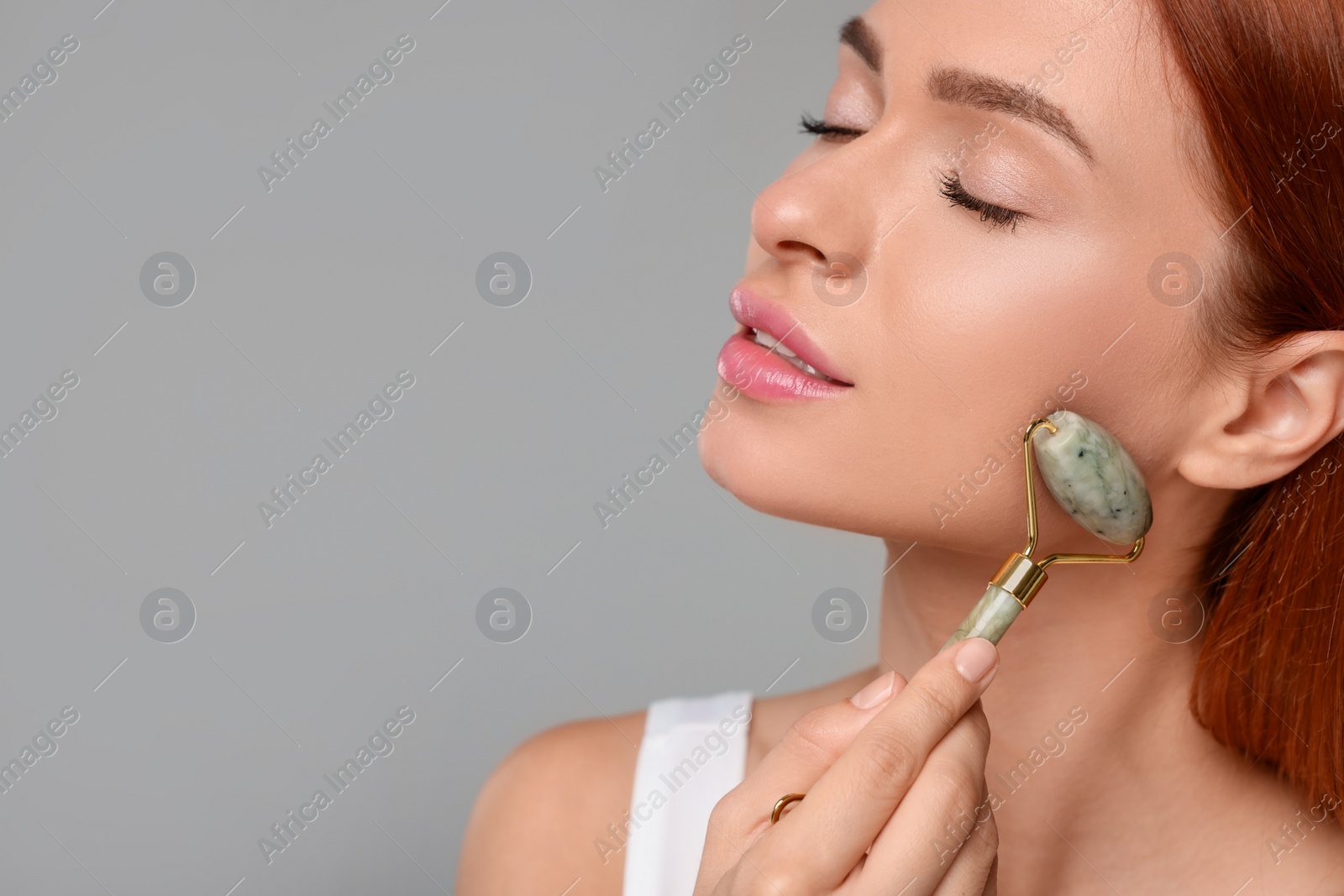 Photo of Young woman massaging her face with jade roller on grey background, closeup. Space for text
