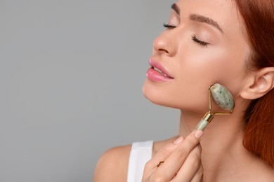 Young woman massaging her face with jade roller on grey background, closeup. Space for text