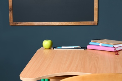 Wooden school desk with stationery and apple near blackboard on grey wall