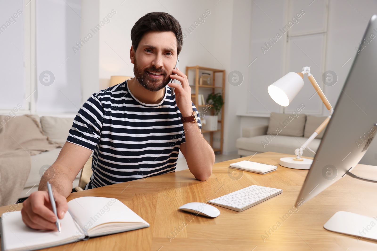 Photo of Home workplace. Happy man taking notes while talking on smartphone at wooden desk in room