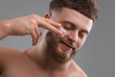 Handsome man applying moisturizing cream onto his face on grey background